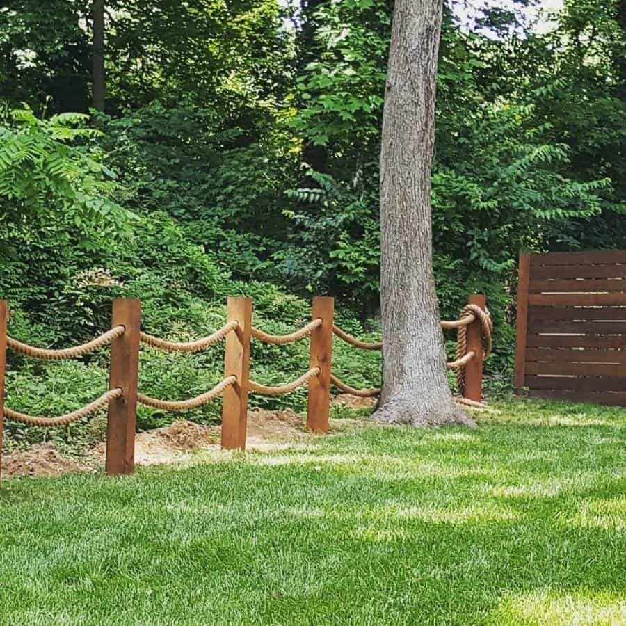 a wooden fence made with rope and short wooden posts encloses a backyard with lush green grass and leafy green trees in the background.  the fence partially goes around a tree.