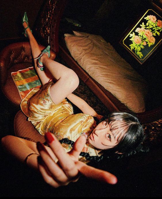 a young woman with black hair and bangs wearing a gold silk chinese dress lays on a brown velvet chaise longue. she is wearing green high heels and has red nail polish. her pose is relaxed, with one leg stretched out and the other bent. she is looking directly at the camera and gesturing with her hand. the setting appears to be a vintage bedroom with dark wood furniture and a floral painting on the wall. the lighting is warm and creates a dramatic, intimate atmosphere.