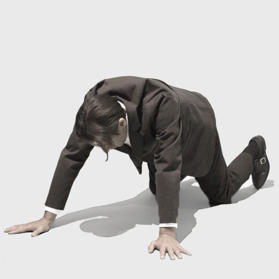 a man in a business suit is kneeling and crawling on a white floor. he is looking down at the floor. his shadow is visible behind him. he is wearing a black suit and black shoes.