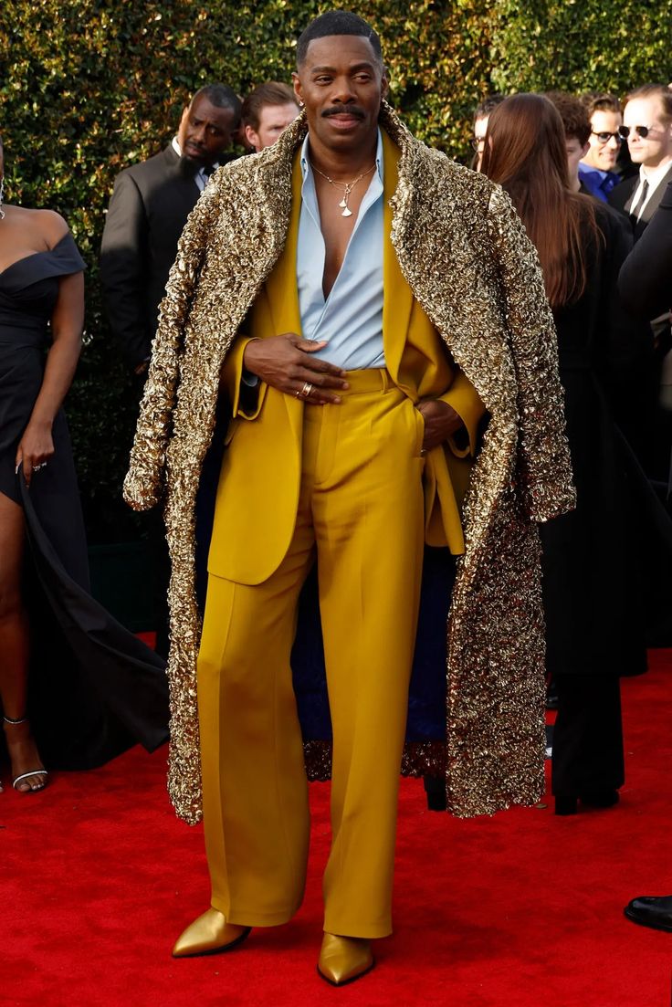 a full shot of a man in a striking mustard yellow suit paired with a luxurious gold sequined coat, standing on a red carpet. he is accessorized with a delicate silver necklace and gold shoes. the background features other attendees dressed in formal wear. the overall ambiance suggests an awards ceremony or a highprofile event. the man's confident posture and stylish outfit make him the focal point of the image.
