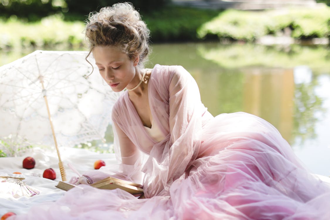 a young woman in a beautiful pink dress is sitting by a lake, reading a book. she is wearing a delicate lace dress and has her hair styled in an elegant updo. a white lace parasol is next to her. the scene is set in a park on a sunny day and is very peaceful.