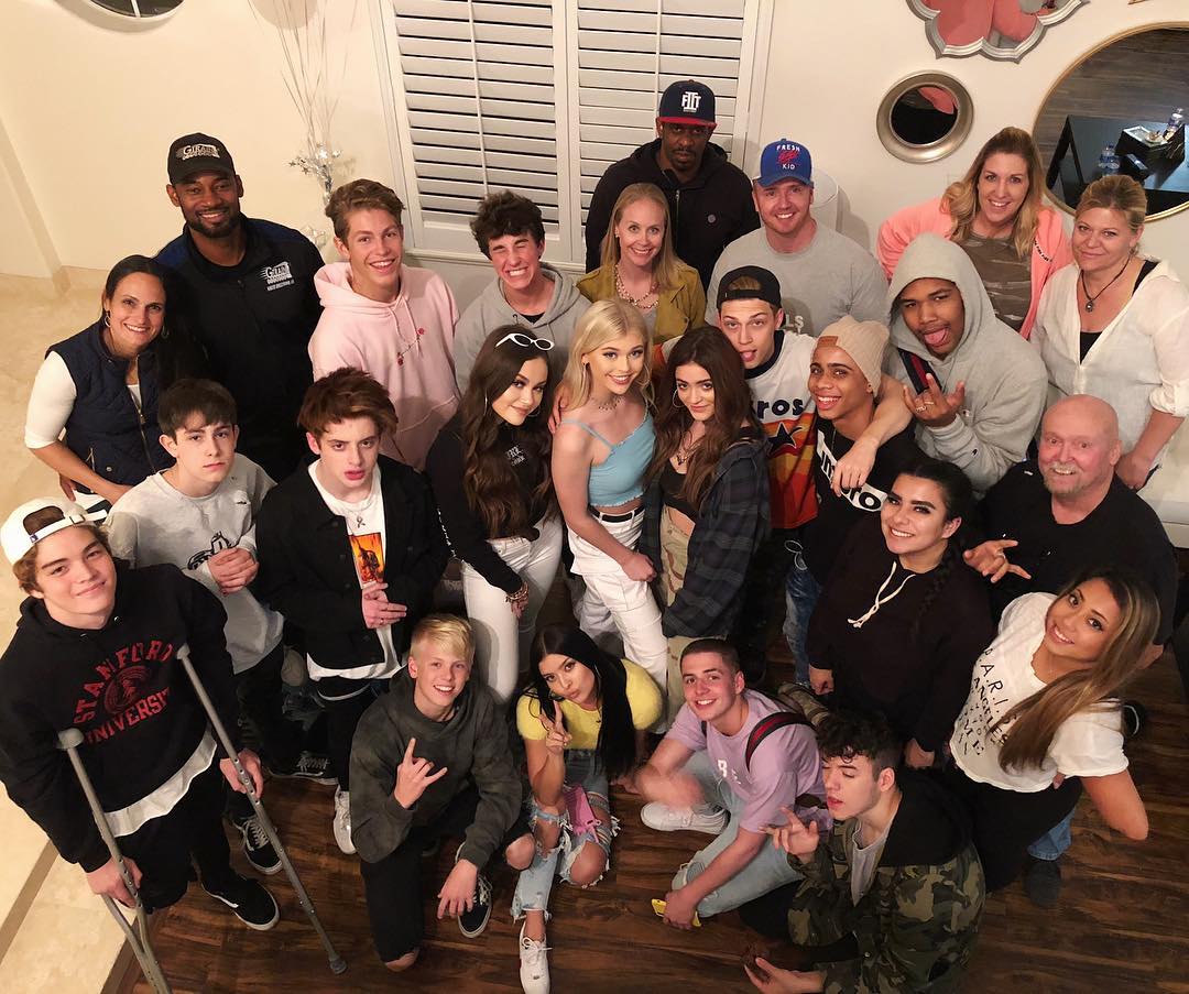 a large group of people pose for a photo indoors. the group consists of adults and teenagers, some standing and some kneeling. one person is using crutches. they are in a room with wood flooring and shuttered windows. there are various items of clothing and accessories visible, including hats, baseball caps, hoodies, and tshirts. the lighting appears to be from indoor sources.