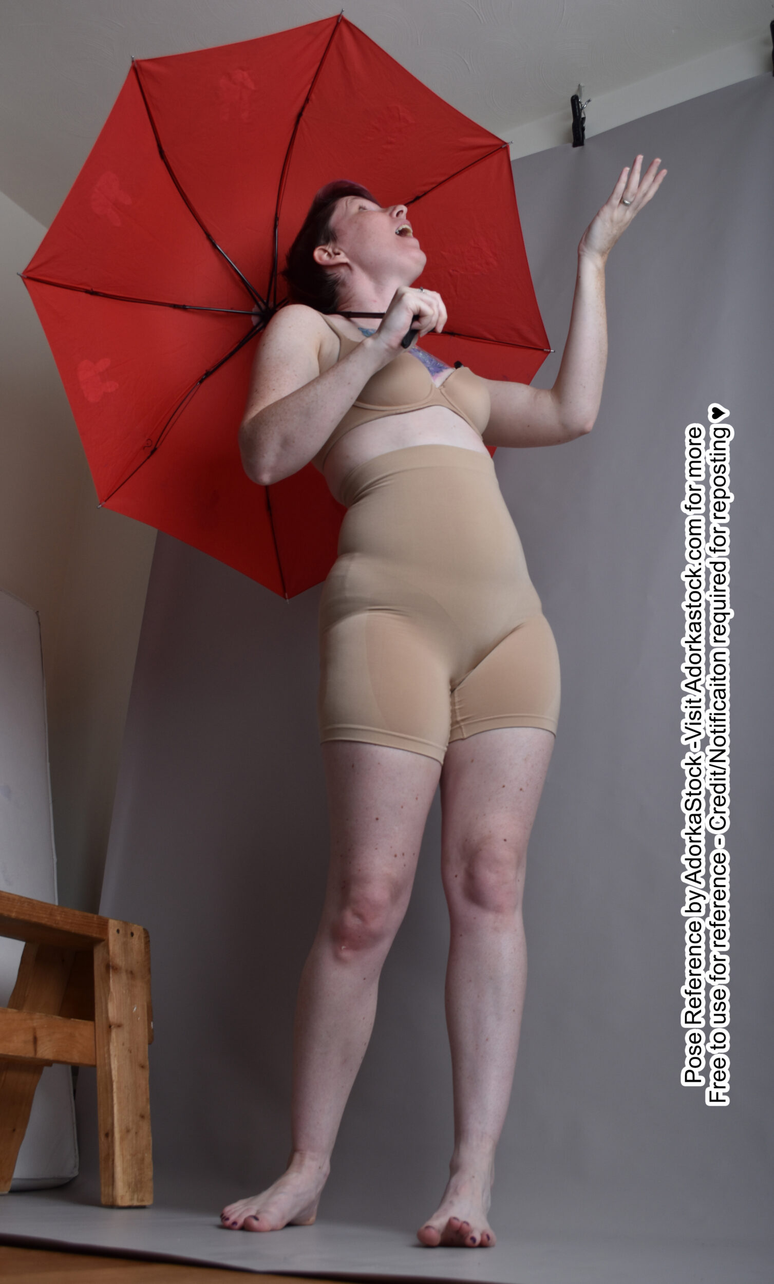 a woman is standing with her right arm raised and holding a red umbrella. she is wearing a highwaisted bodysuit. the woman's right leg is slightly bent, and her left leg is straight. she is facing slightly to her left.  the background is a light gray wall.  this image would be useful as a reference for artists who are drawing or painting the human body.