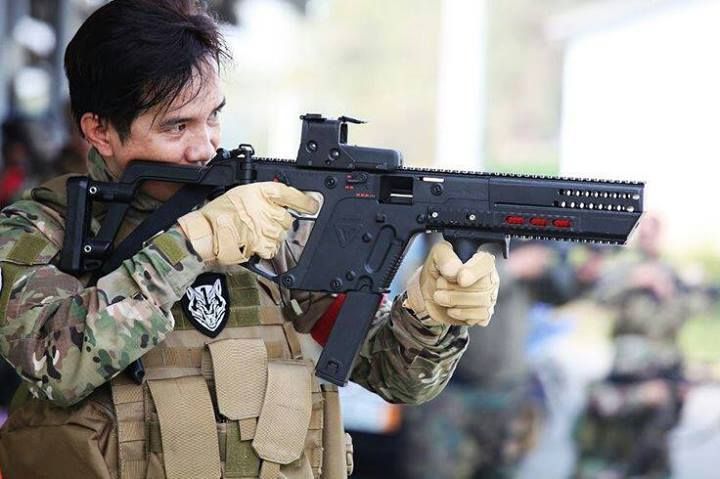 a man in a military uniform, wearing gloves and a tactical vest, is aiming a kriss vector rifle. he has a patch on his uniform. other people in military uniforms are blurred in the background.