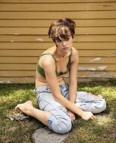 a young woman with short brown hair and blue eyes is sitting in a grassy field. she is wearing a green crop top and blue jeans. she is smiling and looking at the camera. the background is a wooden wall.