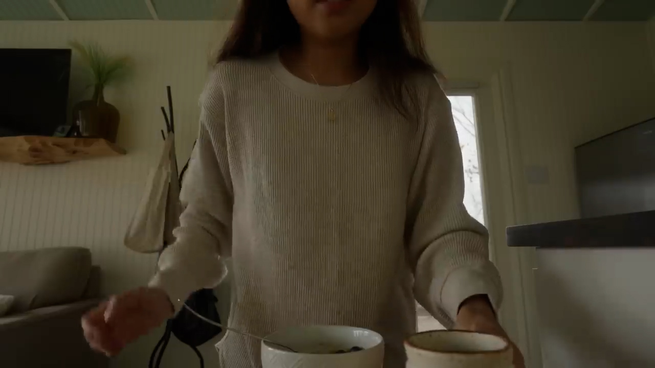 a young woman is seen from the chest down, standing in a softly lit apartment. she holds a bowl of cereal in one hand and a mug in the other. the image is taken from a firstperson perspective, creating a feeling of being present in the moment. the apartment has a cozy and minimalist aesthetic, with neutral tones and natural elements.