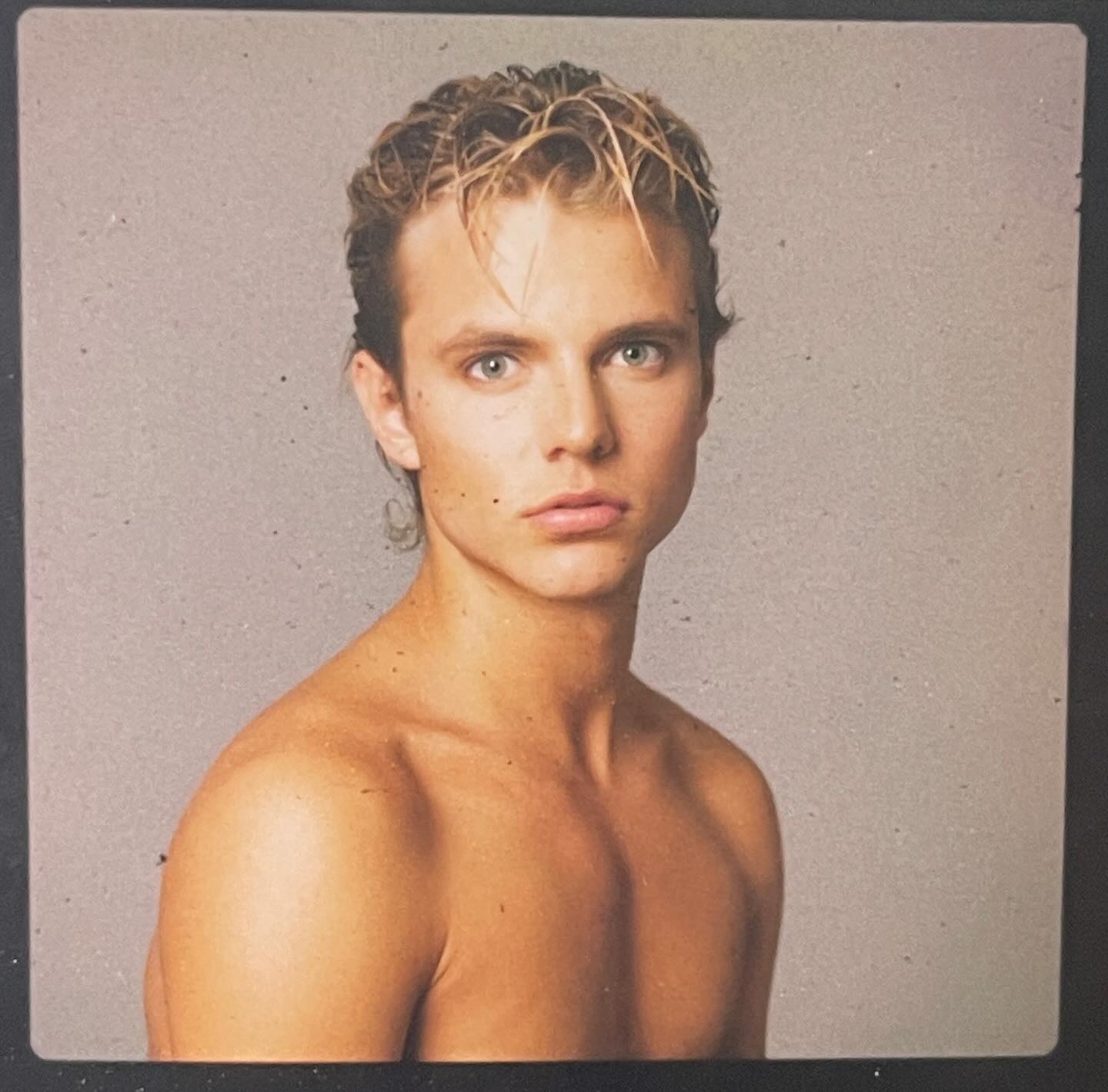 a headshot of a young man with 80s style hair. he is shirtless and looking directly at the camera with a neutral expression.  the background is a solid, muted color and the lighting is soft.