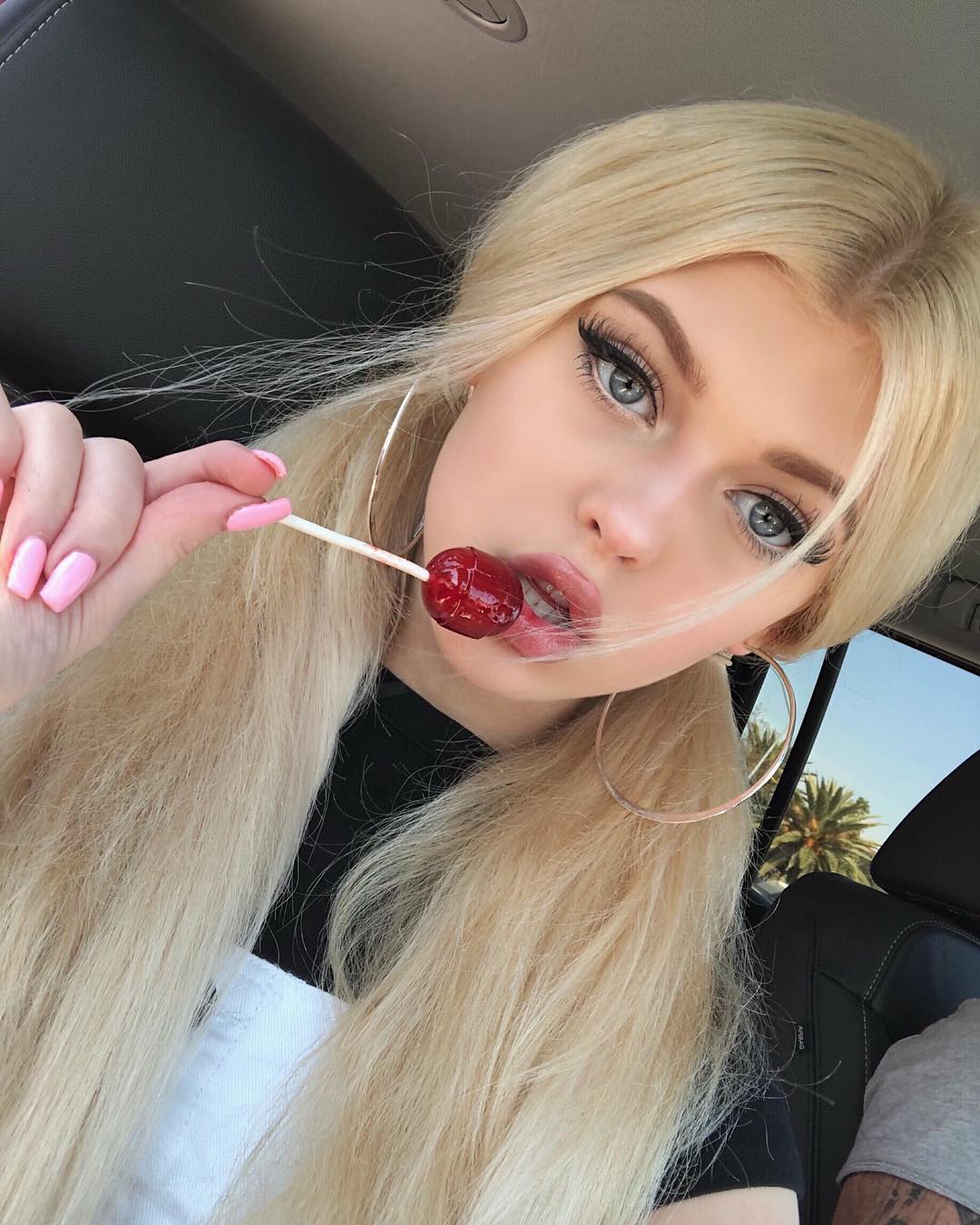 a blonde woman with her hair in pigtails is in a car, holding a red lollipop. she is wearing hoop earrings and has pink nail polish. a palm tree is visible through the car window.