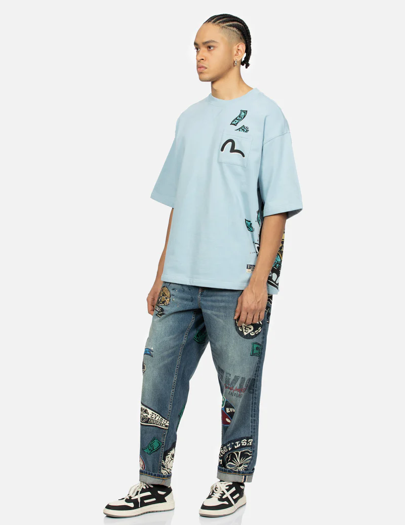 a man is standing against a white background, wearing a light blue oversized comme des garçons play tshirt with a graphic money print and a signature heart logo on the pocket. he's also wearing patched denim jeans with various embroidered patches and white sneakers. the overall style is casual and streetwearinspired.  the image is a studio shot with good lighting.
