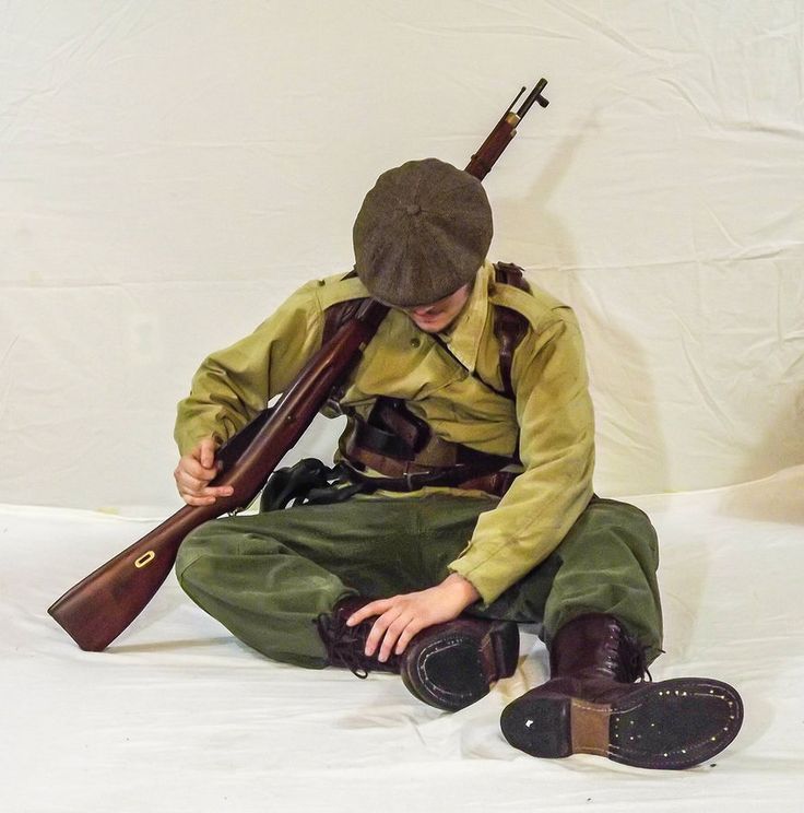 a man dressed in a military uniform sits with a rifle resting on his shoulder. he is looking down and seems to be in deep thought. the man is wearing a green uniform with a brown belt and a brown cap. he is wearing brown boots with black laces. he is holding the rifle with his right hand. he is sitting on a white surface.
