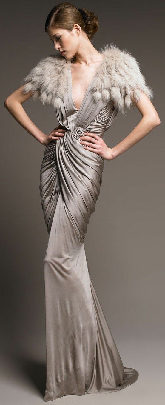 a woman wearing a grey silk gown with a fur shrug poses for the camera. she is looking down at the camera with a sultry expression. her gown is draped and gathered, and she has a elegant, glamorous look. the background is a light grey.