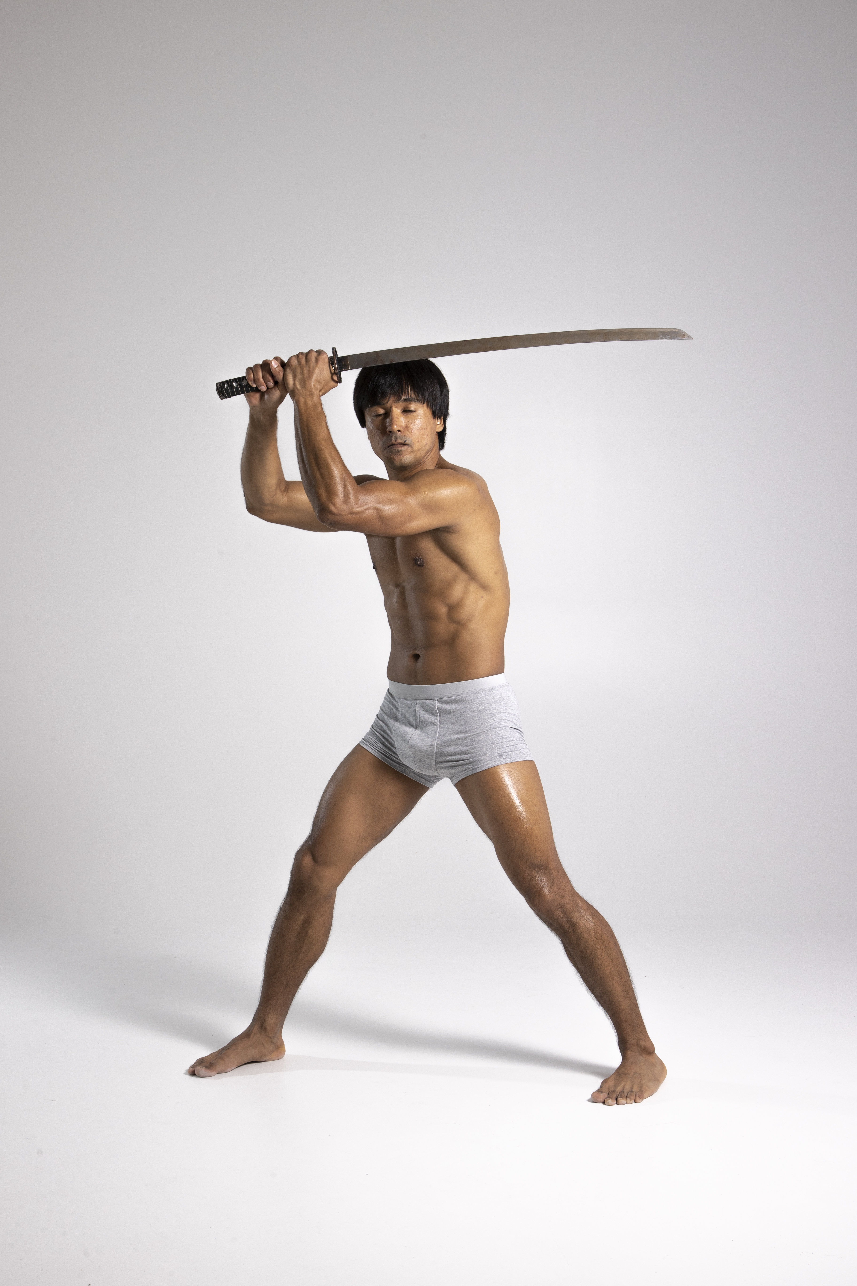 a shirtless, muscular man is shown holding a katana sword over his head in a defensive or attacking pose. he is wearing grey boxer briefs and is standing barefoot on a plain white background. his legs are spread apart for balance.