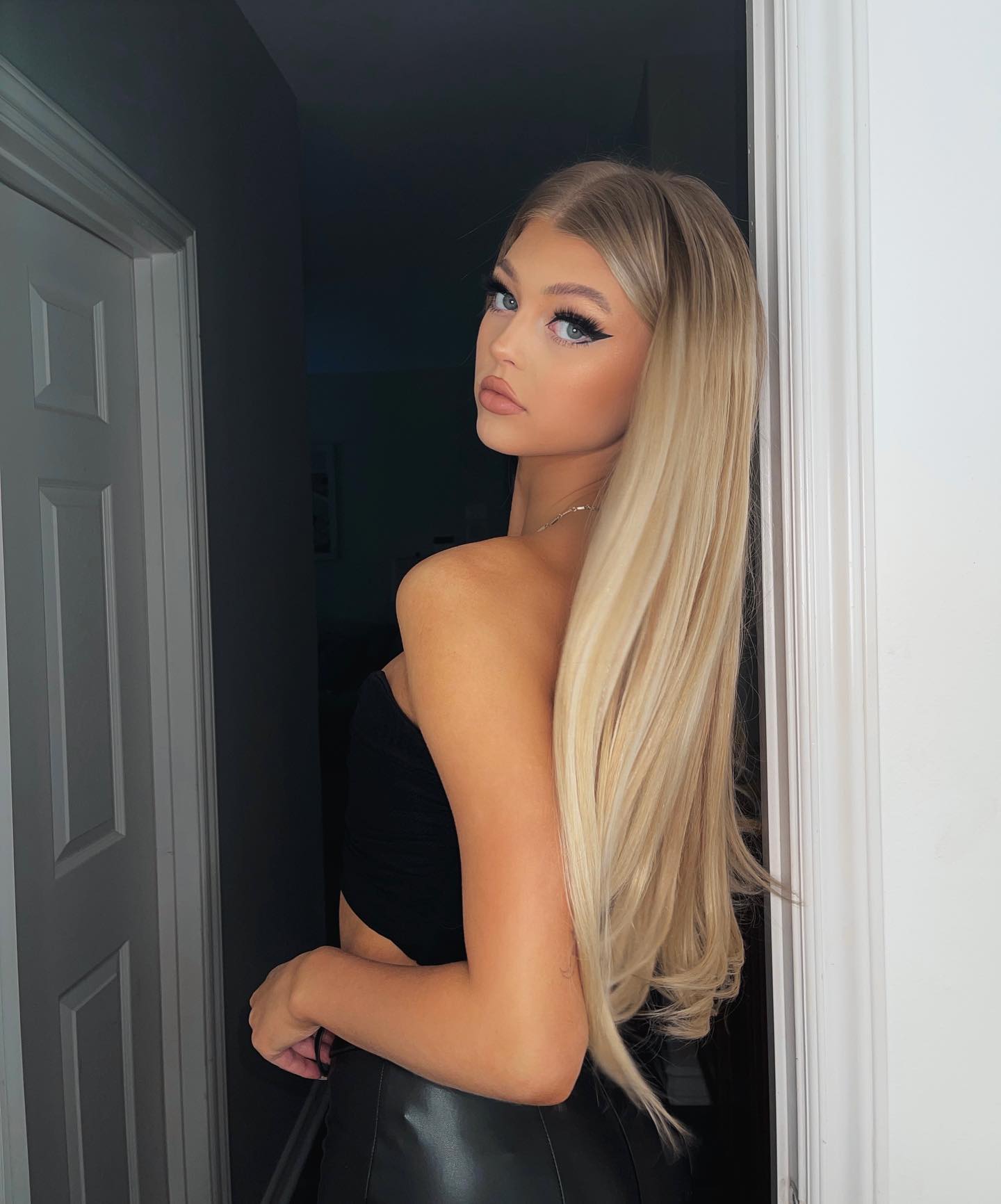 a young woman with long blonde hair, blue eyes, and eyeliner is standing in a doorway. she is wearing a black top and a black leather skirt. she is looking over her shoulder at the camera.