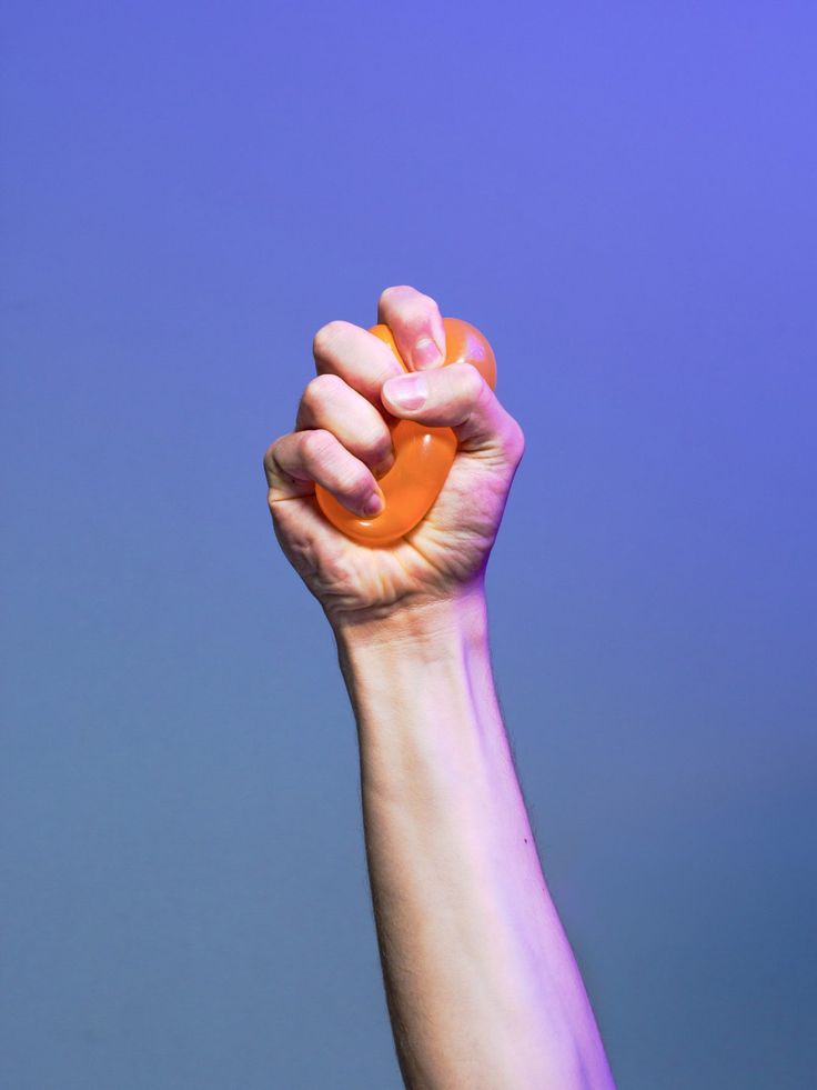a hand squeezing a small orange stress ball with a blue and purple background.