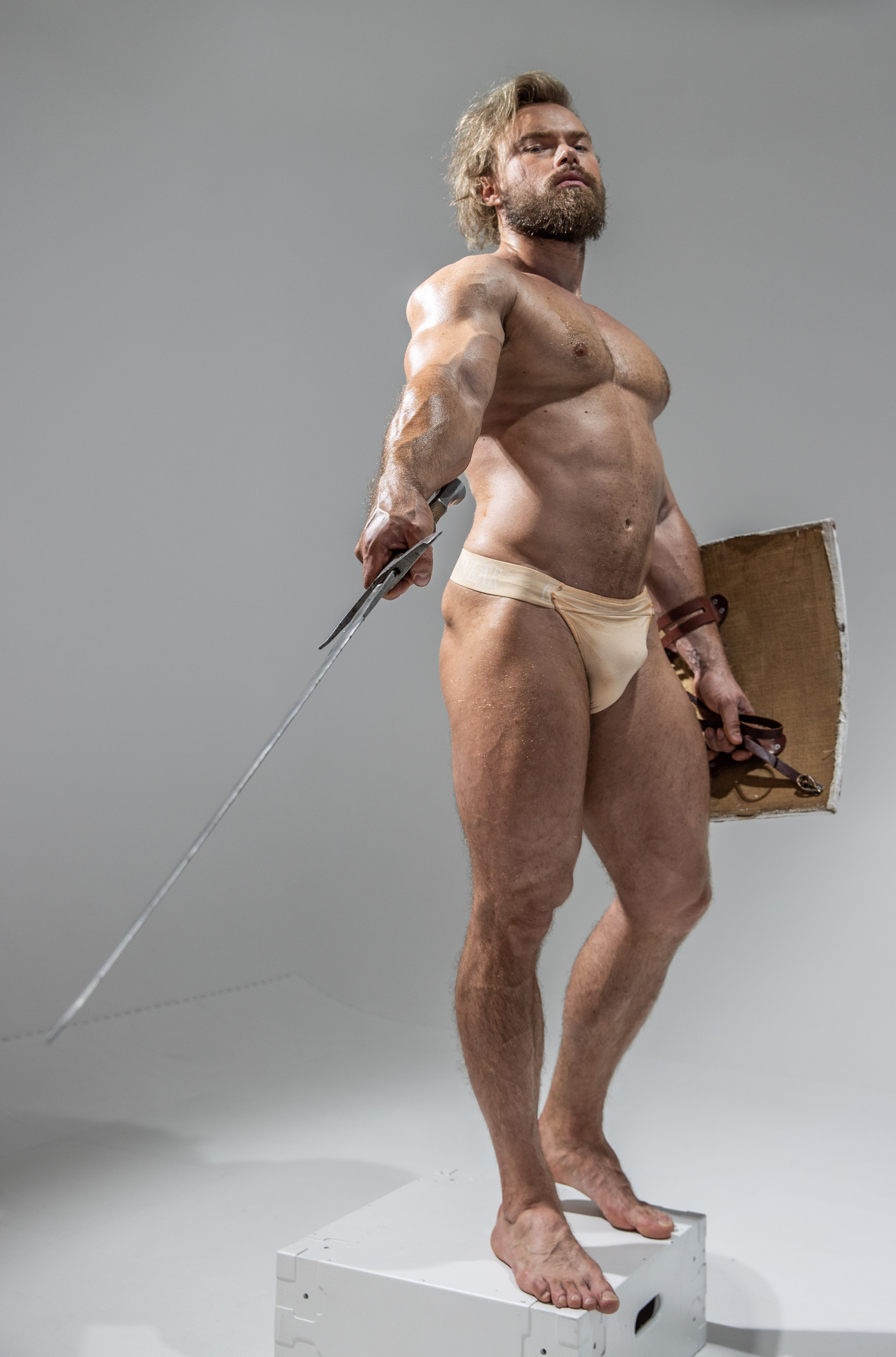a muscular, blonde man stands on a white platform against a white background. he is wearing only beige underwear and is holding a sword and shield. he has a beard and long hair and is looking up and to the right. the image is welllit and shows off the man's physique.