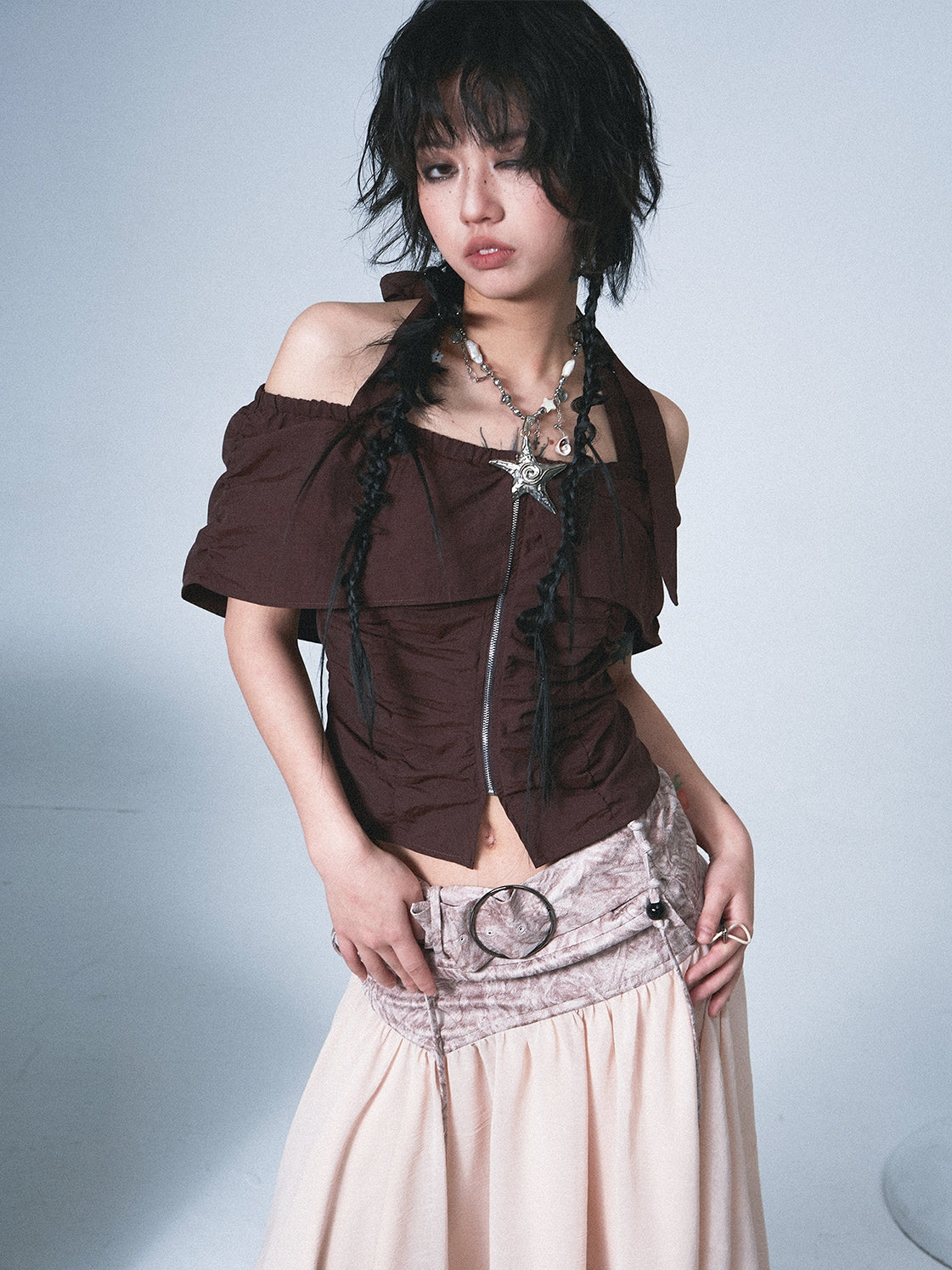a young woman with short, dark hair styled in braids is posing in a studio. she is wearing a brown offtheshoulder top with a ruffled texture and a beige, long skirt. the skirt has a darker beige patterned section at the waist with a large ringlike belt buckle.  she is also wearing a necklace with a starshaped pendant. the overall style is fashionable and slightly bohemian.  the background is a simple, light gray.