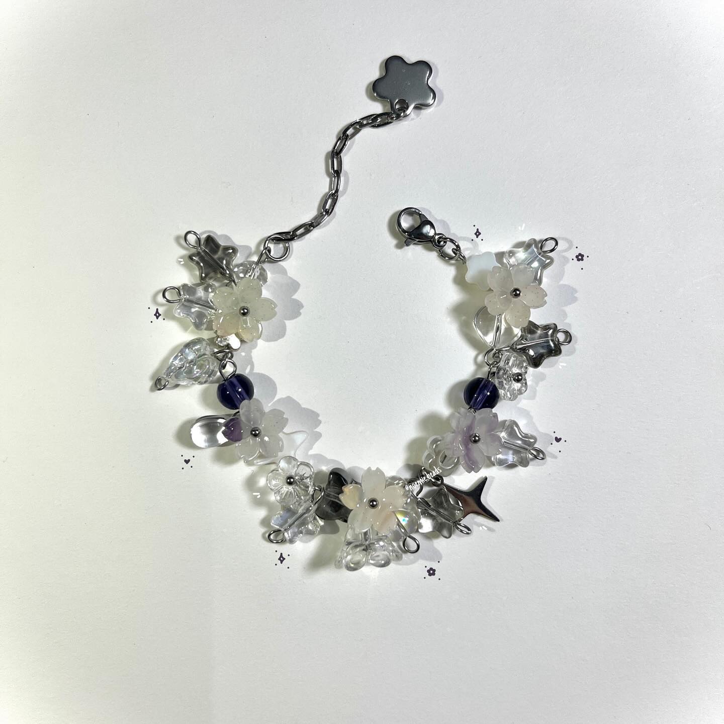 a handmade beaded bracelet featuring white flower beads, clear leaf beads, dark purple round beads, and silver star charms. the bracelet has a silver chain extension and a clasp. the overall style is delicate and whimsical.