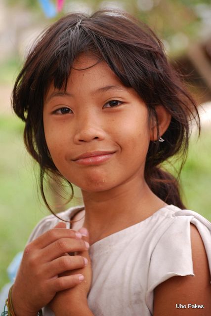 a young asian girl with brown hair is smiling in a portrait. she is wearing a white tshirt and has her hands folded in front of her. the background is blurred and appears to be outdoors. the girl has a small earring in her ear.