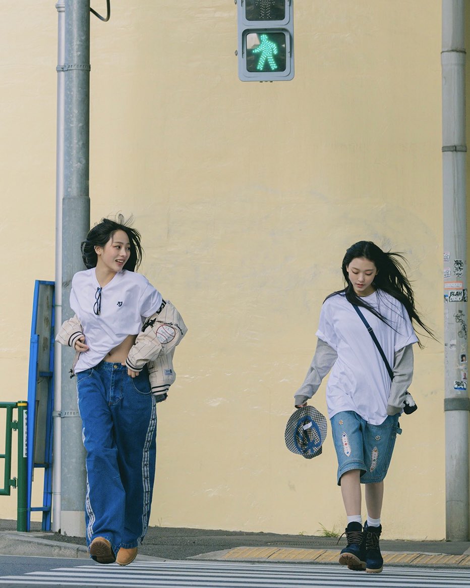 two young women are captured walking across a crosswalk on a sunny day. they exude a casual and trendy street style. the woman on the left sports a white cropped tshirt, baggy blue jeans with white stripes, and brown boots. her friend on the right is dressed in an oversized white tshirt, denim shorts, and black hiking boots. she carries a blue and white hat in her hand. the urban setting and natural lighting enhance the carefree and stylish vibe of the image.