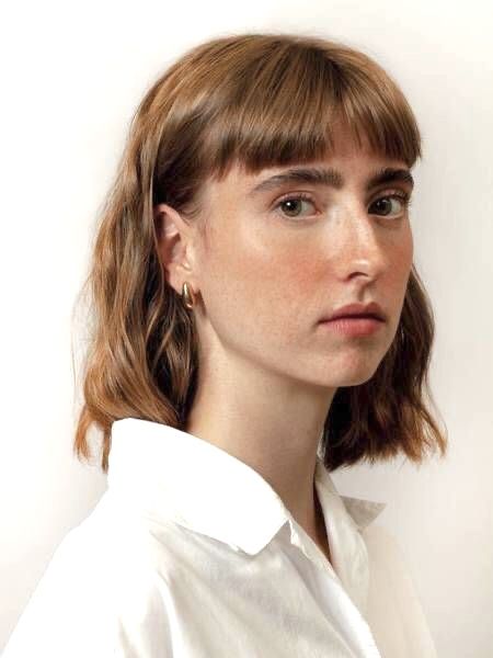 the image is a closeup portrait of a young woman with shoulderlength, wavy brown hair styled with bangs. she has light skin with freckles and is wearing a simple white collared shirt and small gold hoop earrings. her expression is neutral, and the background is a plain, offwhite color. the overall style is minimalist and natural.