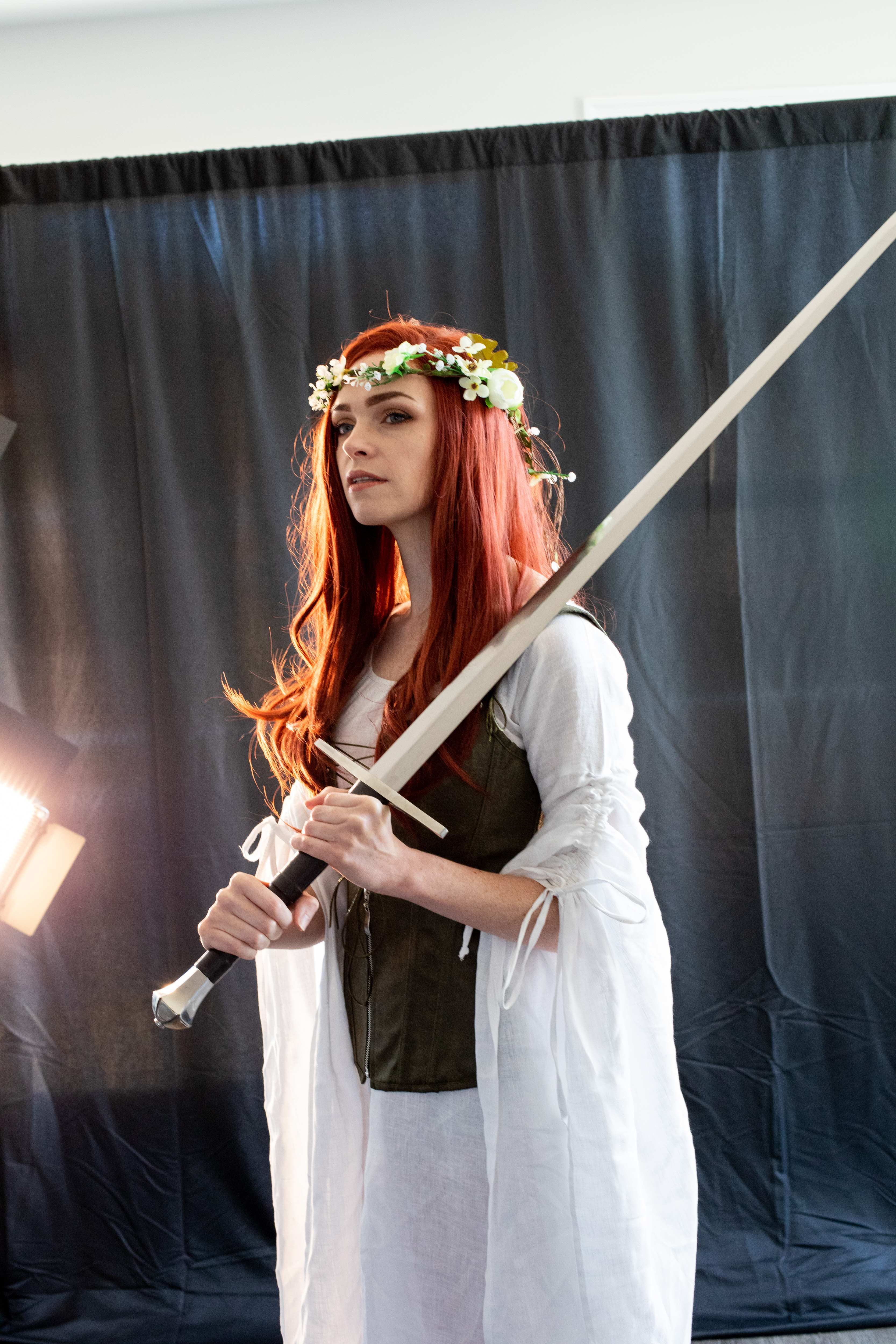 a young woman with long red hair and a flower crown stands against a dark backdrop, holding a sword. she is dressed in a white medievalstyle dress and a green bodice, evoking a warrior or fantasy character. the lighting is soft and diffused, creating a dramatic and ethereal atmosphere.