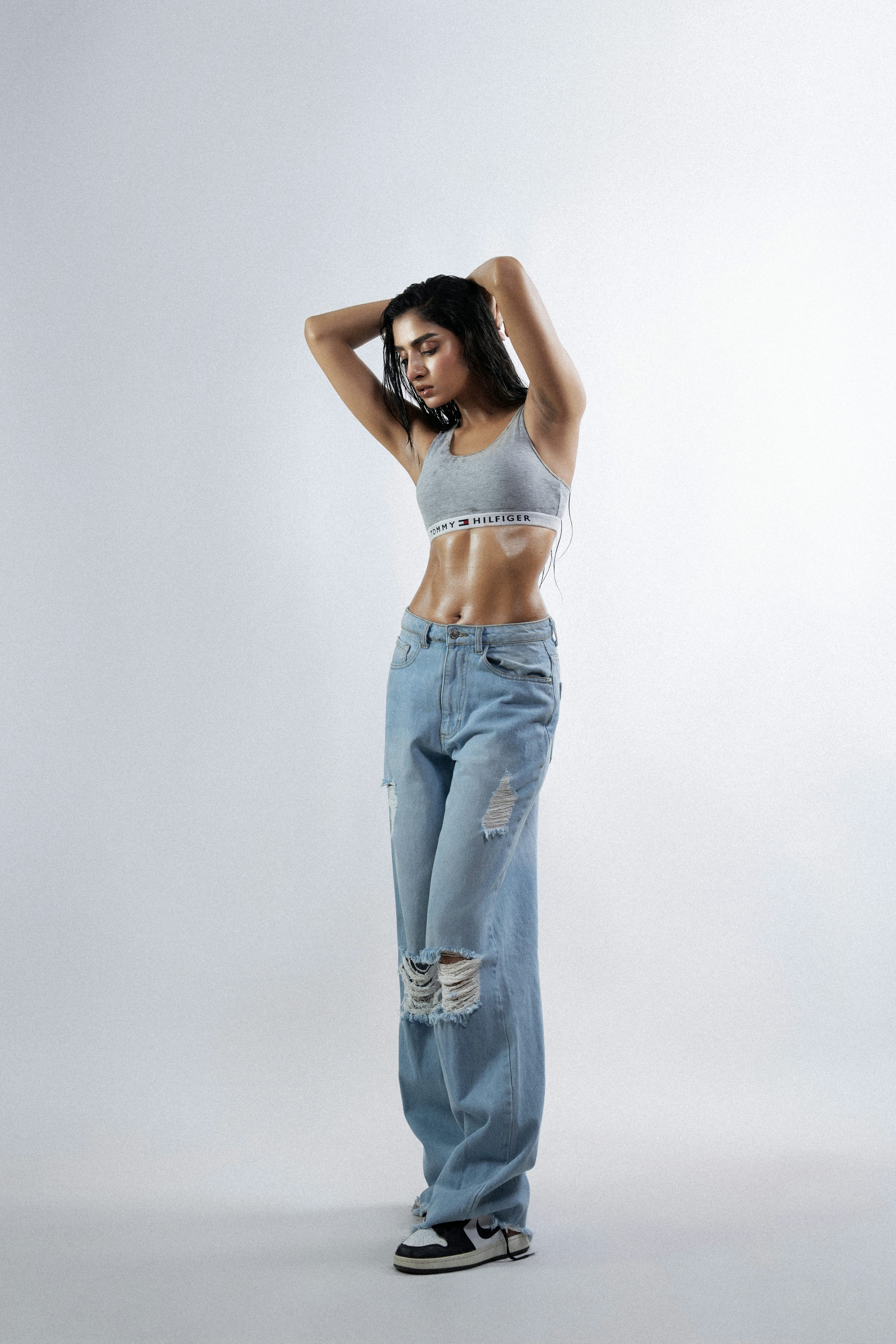 a woman in a gray tommy hilfiger sports bra and ripped light blue jeans stands with her hands behind her head in a studio shot against a white background. she is also wearing black and white sneakers. her abs are visible and she appears to be fit. she is posing in a casual manner.