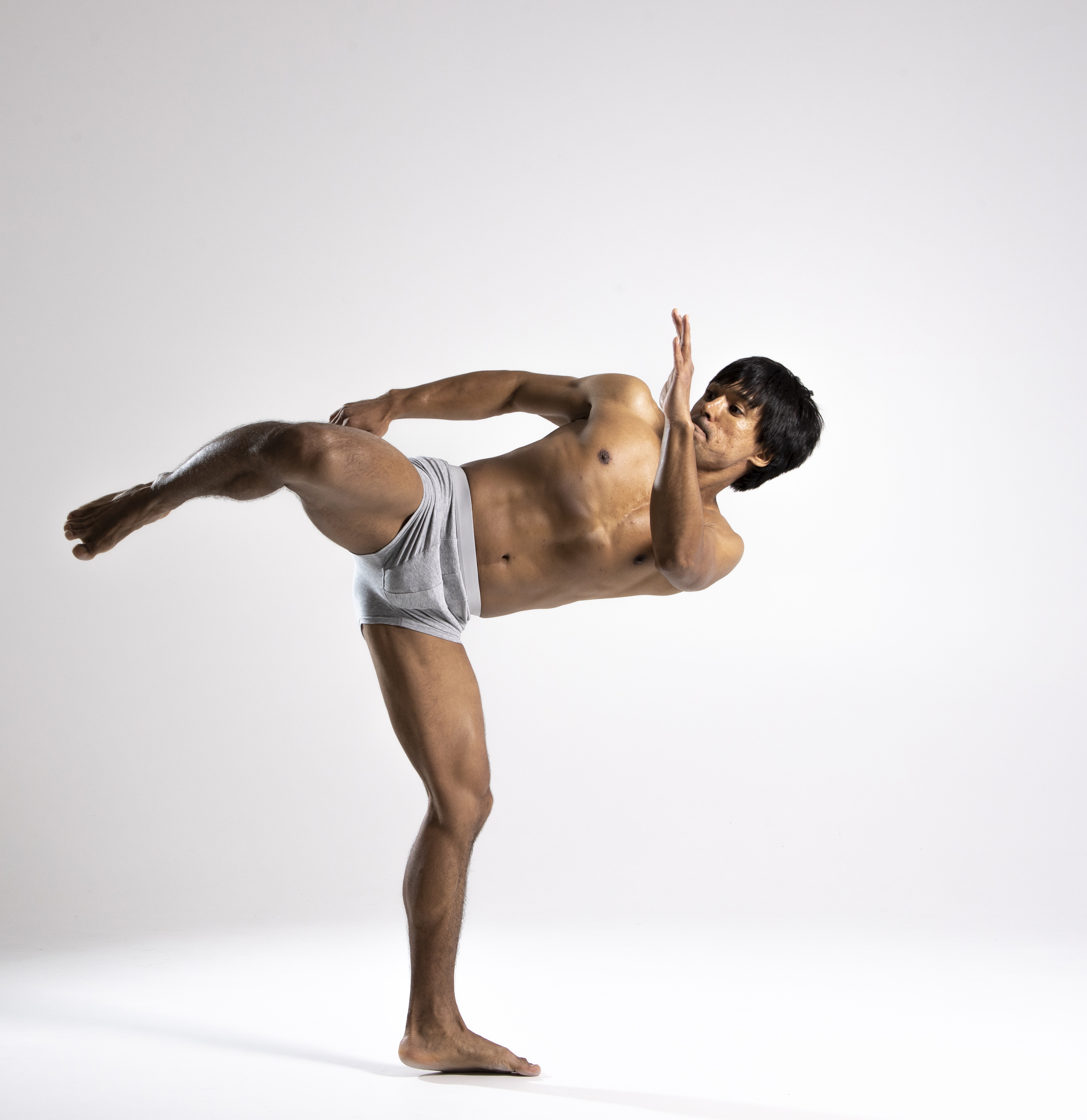 a shirtless, muscular male dancer wearing gray underwear is posing in a studio. he is balancing on one leg, with the other leg raised and extended behind him. his body is arched, and his hand is raised in front of his face. the background is a plain gray.