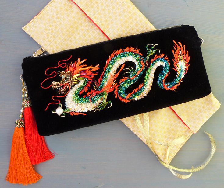 a closeup shot of a black velvet clutch purse adorned with an intricately embroidered dragon. the dragon is made with beads, sequins, and other embellishments, creating a colorful and shimmering effect. the clutch has a red tassel hanging from it. the purse is resting on a yellow fabric with a gold trim.