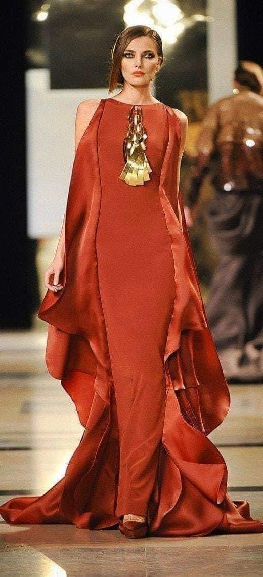 a model walks the runway wearing a stunning terracottacolored gown.  the dress is long and features a flowing, draped design with a high slit. a substantial gold necklace is visible against the dress. the overall aesthetic is elegant and sophisticated, showcasing high fashion. the background is blurred, drawing attention to the model and her dress. the model's makeup is subtle and complements the overall look.