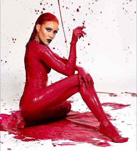 a woman covered in red paint is sitting on a white surface also covered in red paint. she is posing for a studio shot, looking directly at the camera. her entire body is painted, including her hair. she is wearing dark lipstick and her hair appears wet. the scene is illuminated with bright colors.