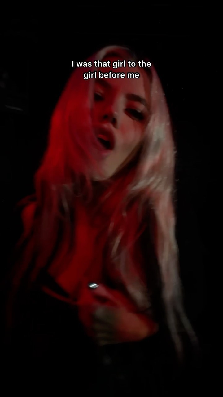a woman with long blonde hair is in the dark with red lighting. the text "i was that girl to the girl before me" is superimposed on the image. she is looking at the camera with her mouth slightly open and is wearing lipstick.