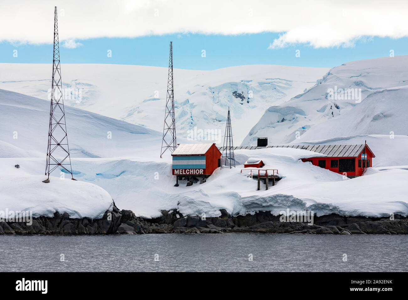 a red and white building stands out against the white snow and ice of antarctica. the building is identified as "melchior" and is likely a research base or outpost. the mountains and ocean in the distance are covered in snow and ice, creating a starkly beautiful and remote landscape.