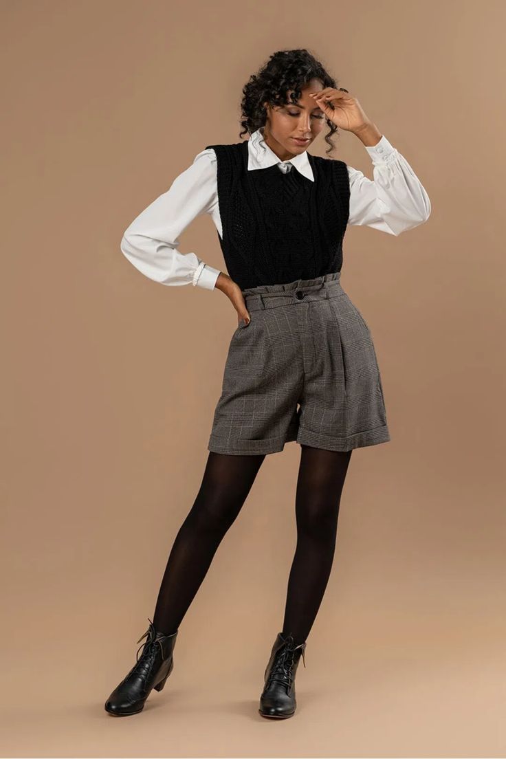 a fulllength studio shot of a young woman, showcasing a fashionable autumnal outfit.  she's wearing a black cable knit sweater vest over a white collared shirt, paired with highwaisted grey plaid shorts and black tights. her footwear consists of black laceup ankle boots. the model is posing with one hand on her forehead, suggesting a slight pensive or thoughtful expression.  the background is a neutral, light brown tone.