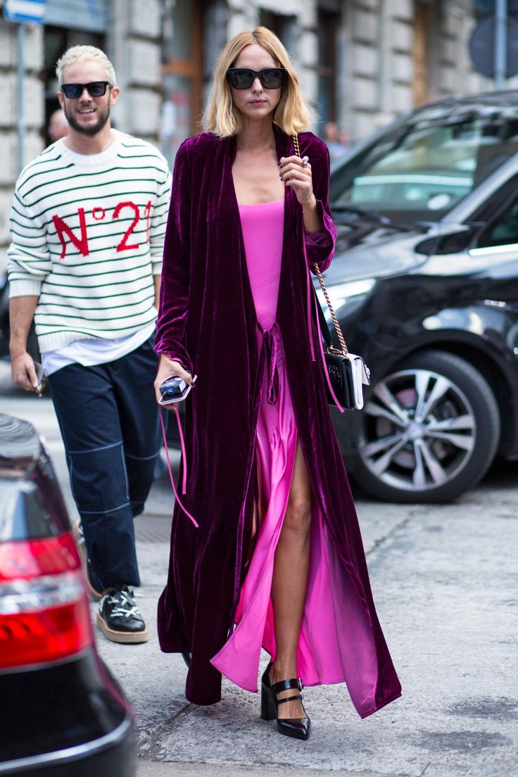 a stylish woman in a long purple velvet coat over a pink dress is walking down a city street with a man. the woman is wearing sunglasses and heels, and carrying a handbag. the man is wearing a striped sweater and pants. they appear to be a couple and are dressed in fashionable attire. the scene is set during the daytime, and the background includes buildings and parked cars.