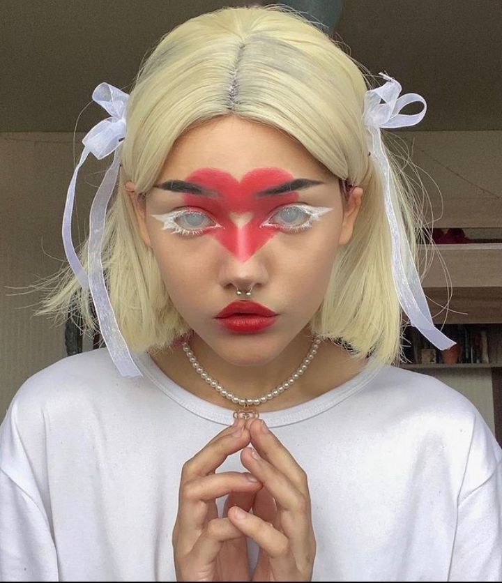 a young woman with blonde hair, white wig, red heartshaped makeup, red lipstick, white shirt, white bow, pearl necklace, nose ring, white eyes and looking at the camera.
