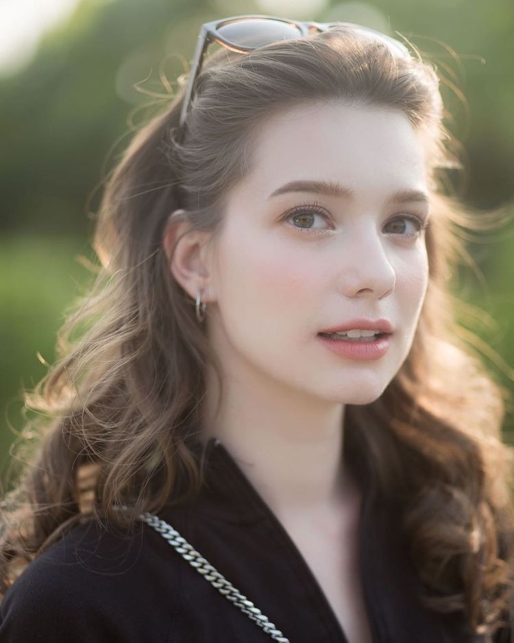 closeup portrait of a young, attractive woman with long, wavy brown hair. she's wearing sunglasses perched on her head, a simple black jacket, and subtle jewelry (earrings and a necklace). the background is softly blurred, showcasing a natural outdoor setting with green foliage. the lighting suggests it's daytime, with sunlight illuminating her face and hair. the overall mood is serene and elegant.