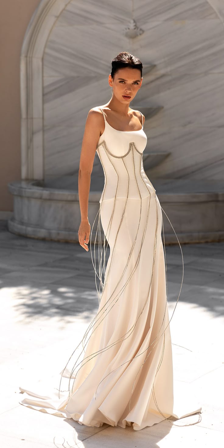 a woman is standing outside, posing in a strapless white corset dress. the dress has spaghetti straps, and decorative silver strands hanging down. she is in front of a marble building with an ornate water fountain.