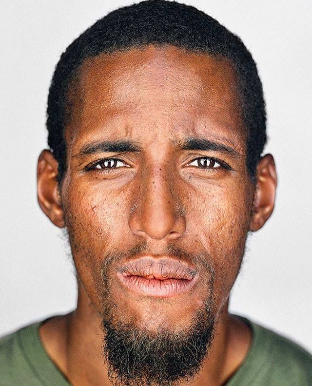a closeup portrait of a man with dark skin, a short beard, and closely cropped hair. he is looking directly at the camera with a serious or slightly concerned expression. the background is plain and neutral.