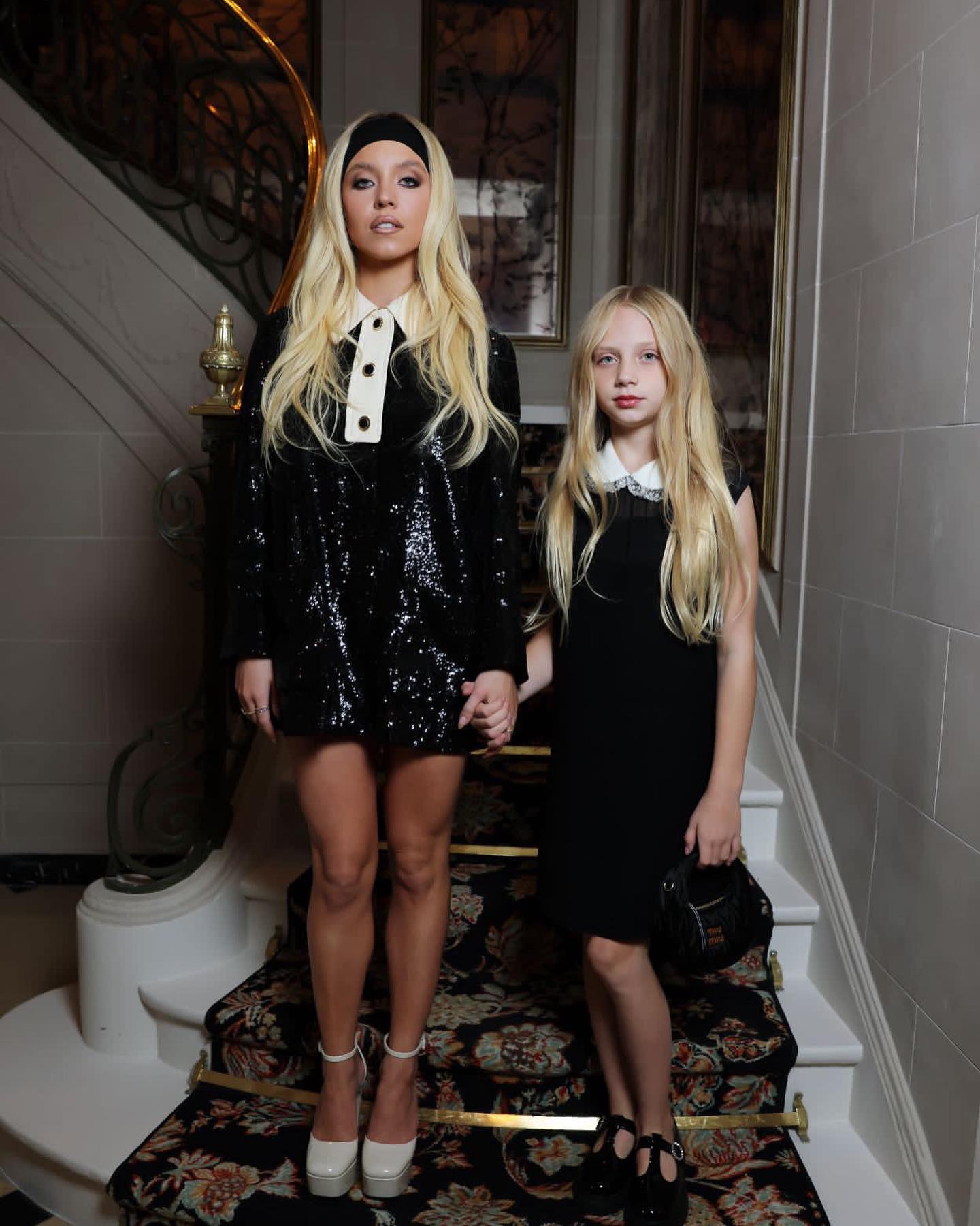 sydney sweeney is standing on a staircase next to a young girl, both wearing black dresses with white collars. sydney is wearing platform heels and a headband. they are holding hands.