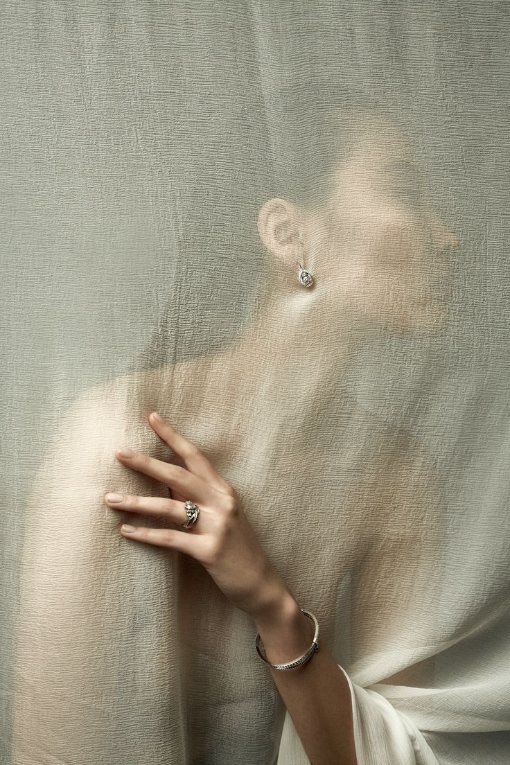 the image is a fashion photograph showcasing a woman's jewelry. the model's face and upper body are partially obscured by a sheer, lightbeige fabric draped artistically around her. only her hand and part of her face are visible.  she is wearing a silver ring, silver earrings, and a silver bracelet. the focus is on the jewelry's design and the overall mood, which is elegant, mysterious, and sophisticated.  the sheer fabric creates a sense of both intimacy and mystery, highlighting the delicate beauty of the accessories. the background is a consistent textured fabric creating a soft, muted backdrop.