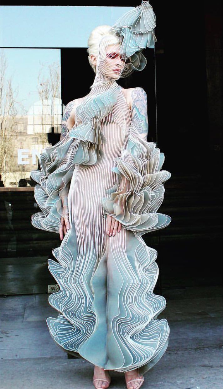 a woman in a white futuristic dress with an intricate, abstract, and wavy design, she is posing on a runway. the design is unique and creative, giving an artistic and elegant look.  the model has a beautiful and stylish look with a unique pose.