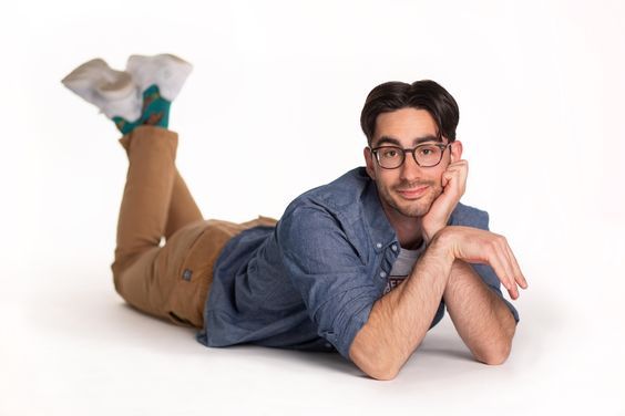 a man with glasses is laying on the floor and smiling. he has on a blue shirt and brown pants. his hand is resting on his cheek. the background is white.