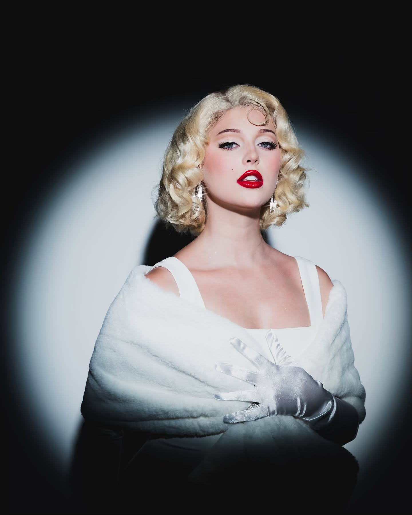 a woman dressed as marilyn monroe is shown in a portraitstyle image. she has blonde, styled hair with a prominent curl on her forehead. her makeup includes bold red lipstick and defined eyebrows. she wears a white dress and a white fur shawl, and her hands are covered in white gloves. she is accessorized with dangling earrings. a spotlight illuminates her against a dark background. the overall look is a classic hollywood glamour impersonation.