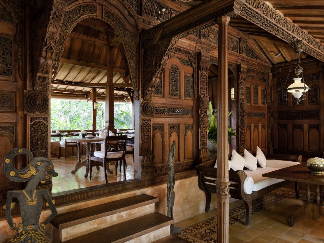 this image shows the interior of a luxurious home. it has intricate wood carvings adorning the walls and pillars. there are two distinct areas: a living room with a couch and coffee table in the foreground, and a dining area with tables and chairs in the background. natural light streams in from the outdoors through the open walls. the color palette is dominated by shades of brown and beige, reflecting the extensive use of wood in the construction.