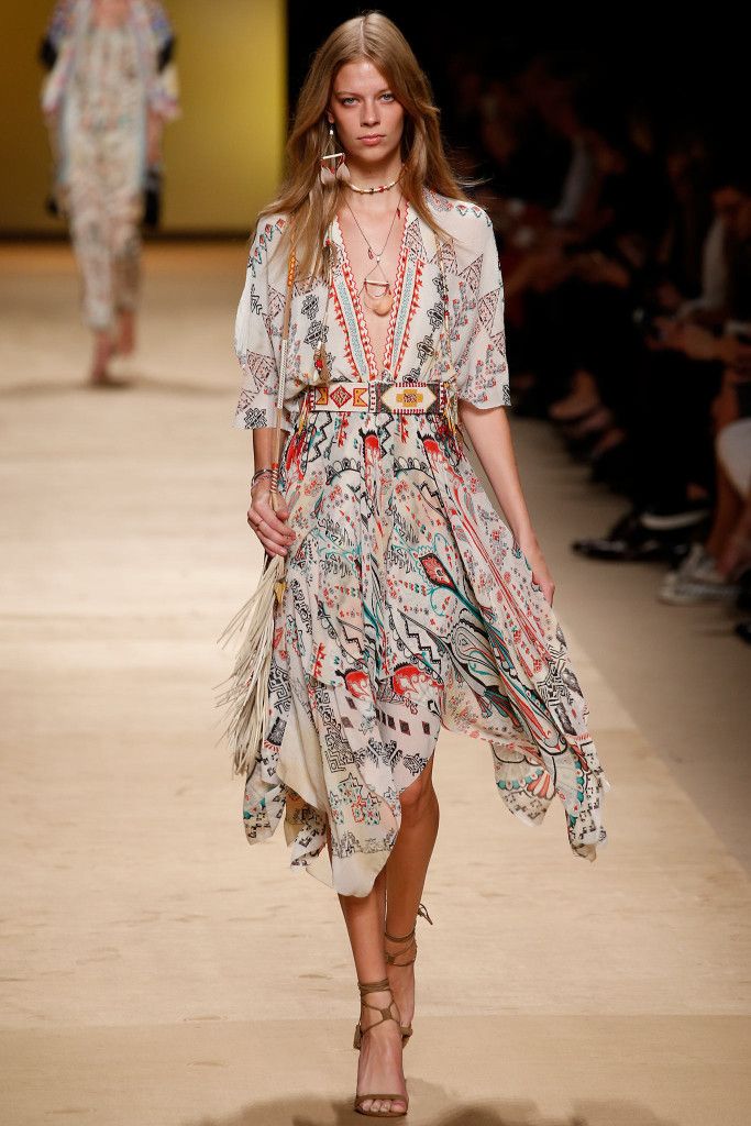 a model walks the runway wearing a bohemianstyle dress. the dress is longsleeved and features a multicolored print on a beige background.  the model also wears several accessories, including a belt with ethnic patterns, necklaces, earrings and sandals. she carries a fringed handbag. the setting appears to be a fashion show.