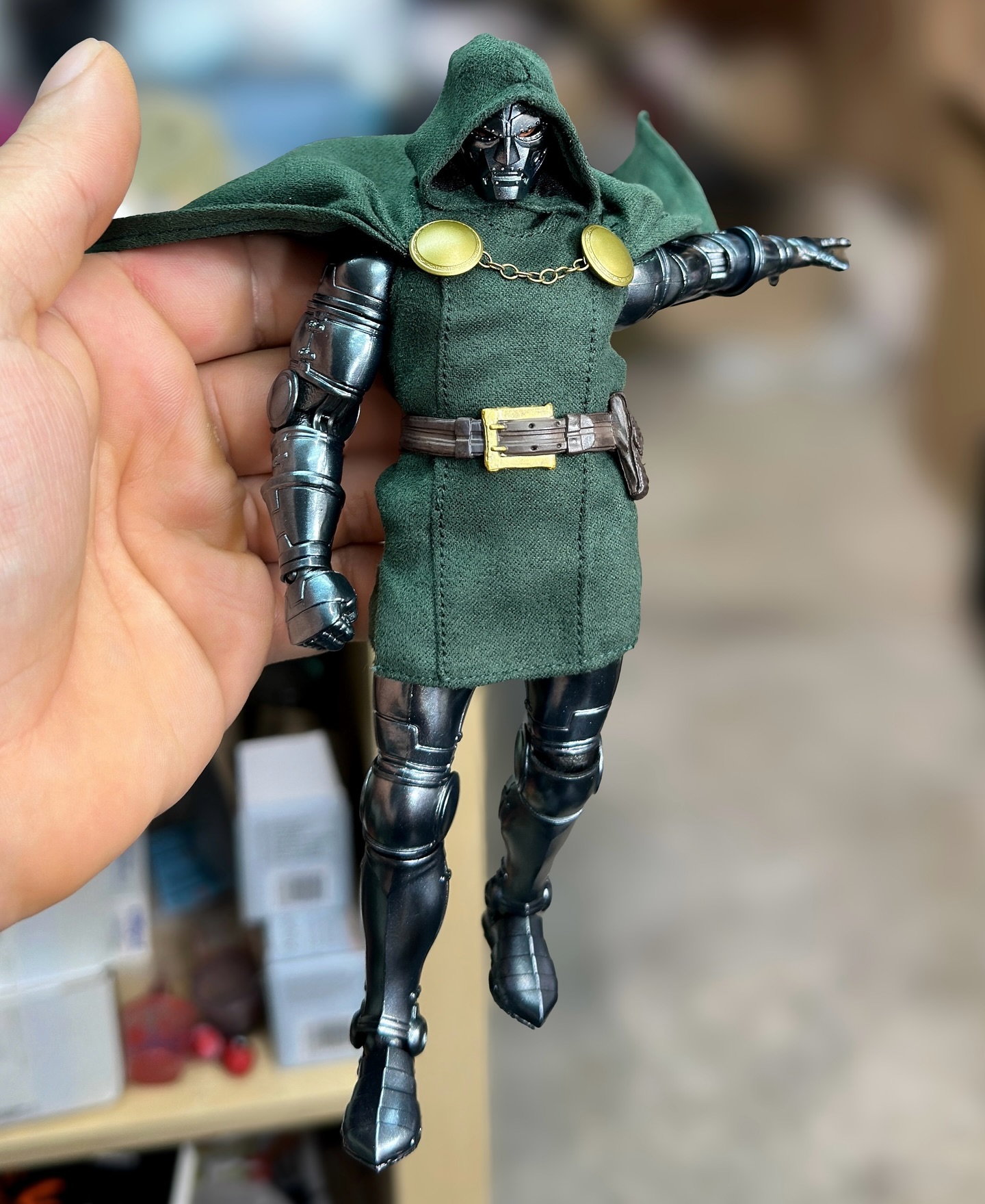 a hand is holding a doctor doom action figure. the figure has a metal body, a green cloak with a hood, a belt, and gold detailing. the background is blurry.