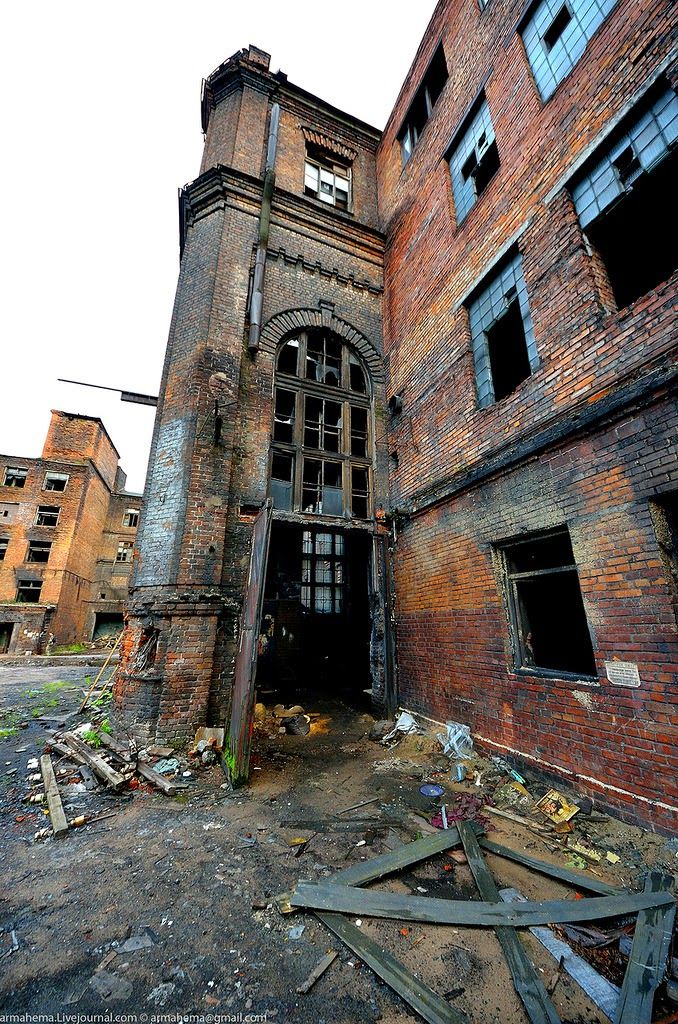 the image shows a dilapidated brick building, likely an abandoned factory, with significant fire damage and decay. the brickwork is dark and sootstained, and many windows are broken.  the ground is covered in rubble, debris, and litter, indicating longterm neglect.  the architecture of the building suggests an industrial past, possibly from the early to mid20th century.  the overall mood is one of desolation and decay. a section of a neighboring, similarly aged and damaged building is visible in the background. the lighting is dim and overcast, enhancing the somber atmosphere.
