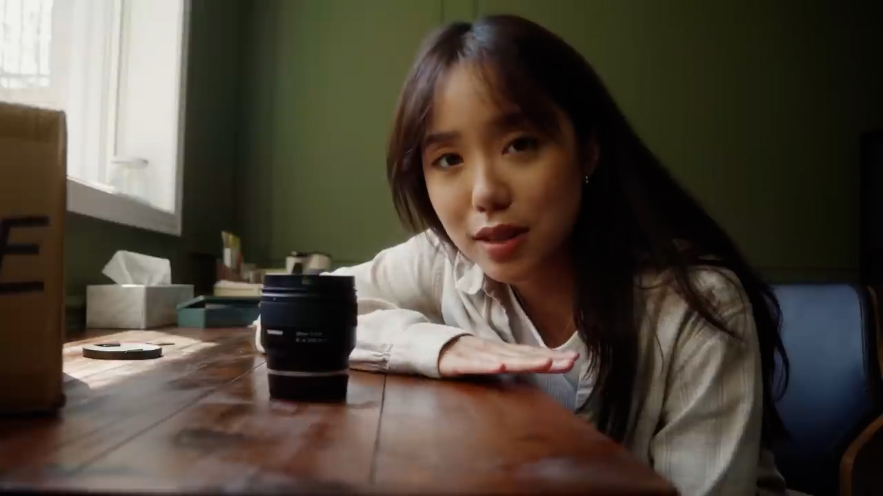 a young woman sits at a desk in a softly lit room, gesturing towards a camera lens in front of her. she appears to be filming a video, potentially a review or unboxing. the image suggests a comfortable and approachable atmosphere.