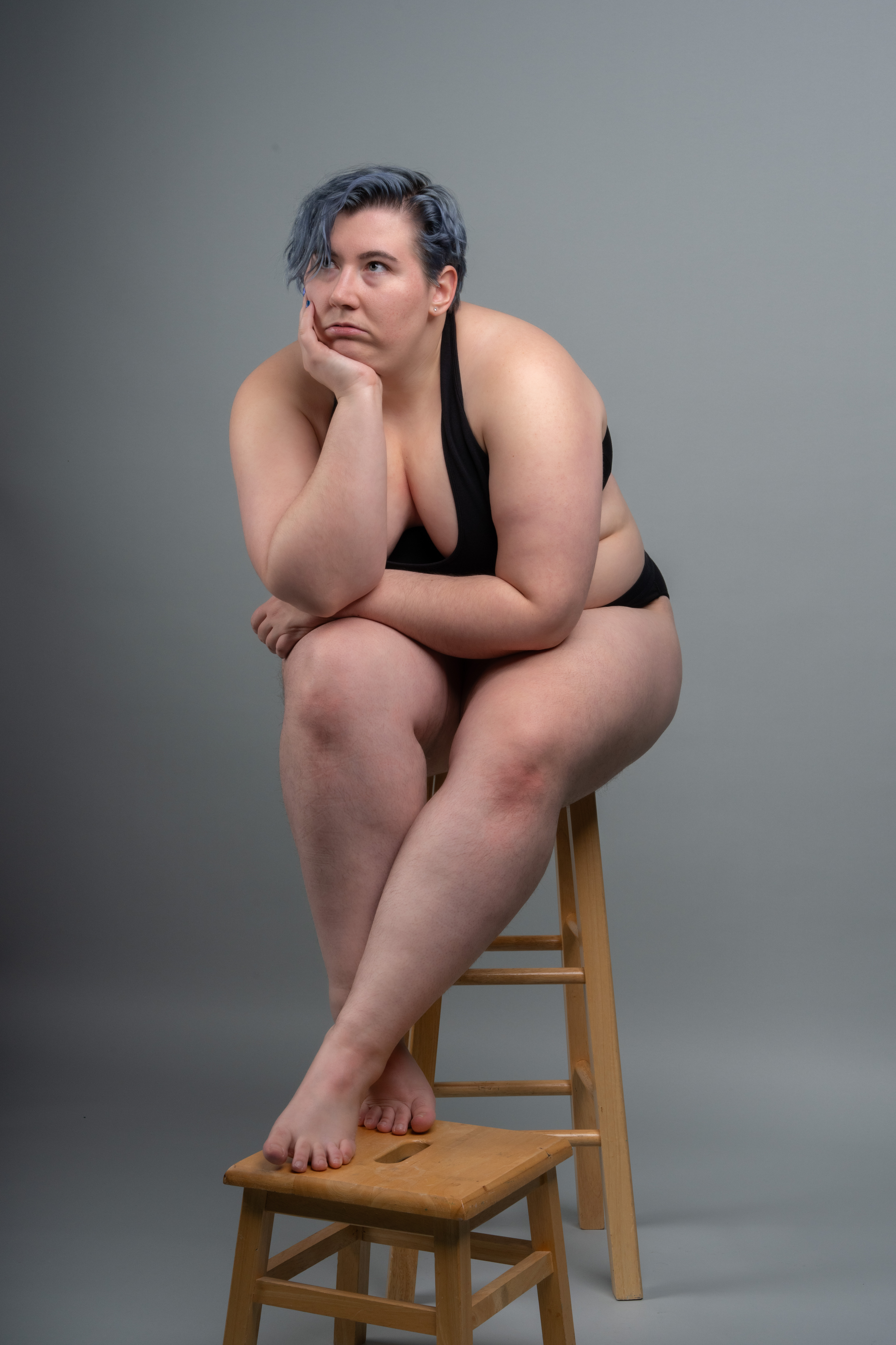 a plussize woman with blue hair is sitting on a wooden stool in a studio setting. she is wearing black underwear and has her legs crossed. she appears to be deep in thought, resting her head on her hand. the background is a plain gray color.