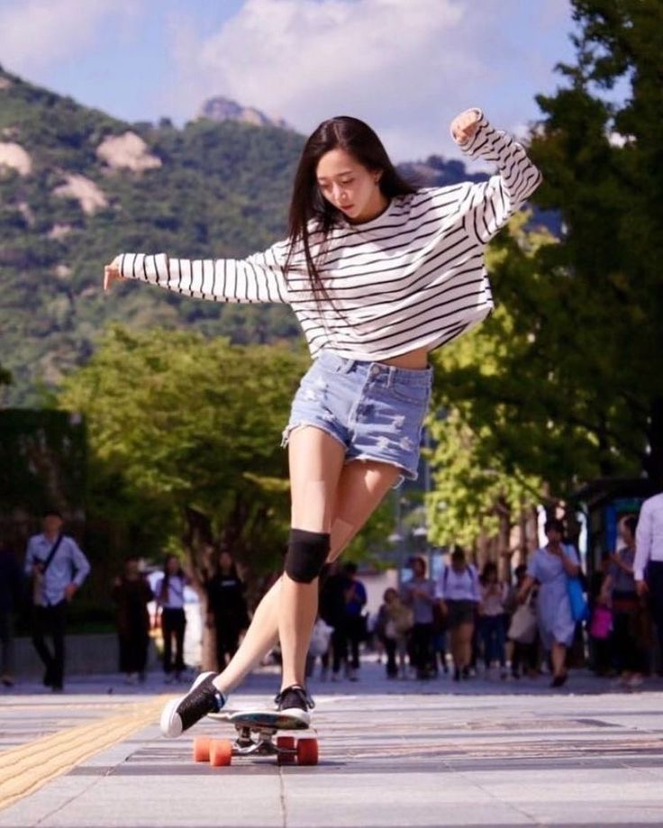 the image shows a young asian woman skateboarding down a city street. she's wearing a striped longsleeve shirt, denim shorts, and knee pads.  she's gracefully balancing on her longboard, looking down at it. the background features mountains and other pedestrians, suggesting a bright, sunny day in an urban setting. the overall impression is one of youthful energy and casual, active fun.